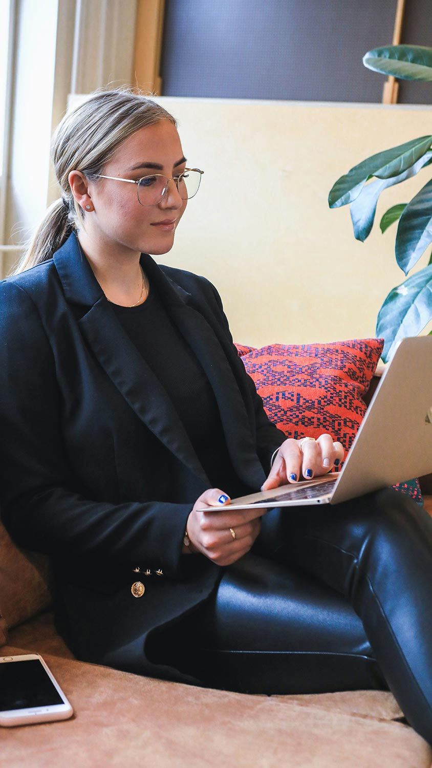 A woman working on a laptop while seated on a couch, focusing on the screen with a smartphone placed beside her.