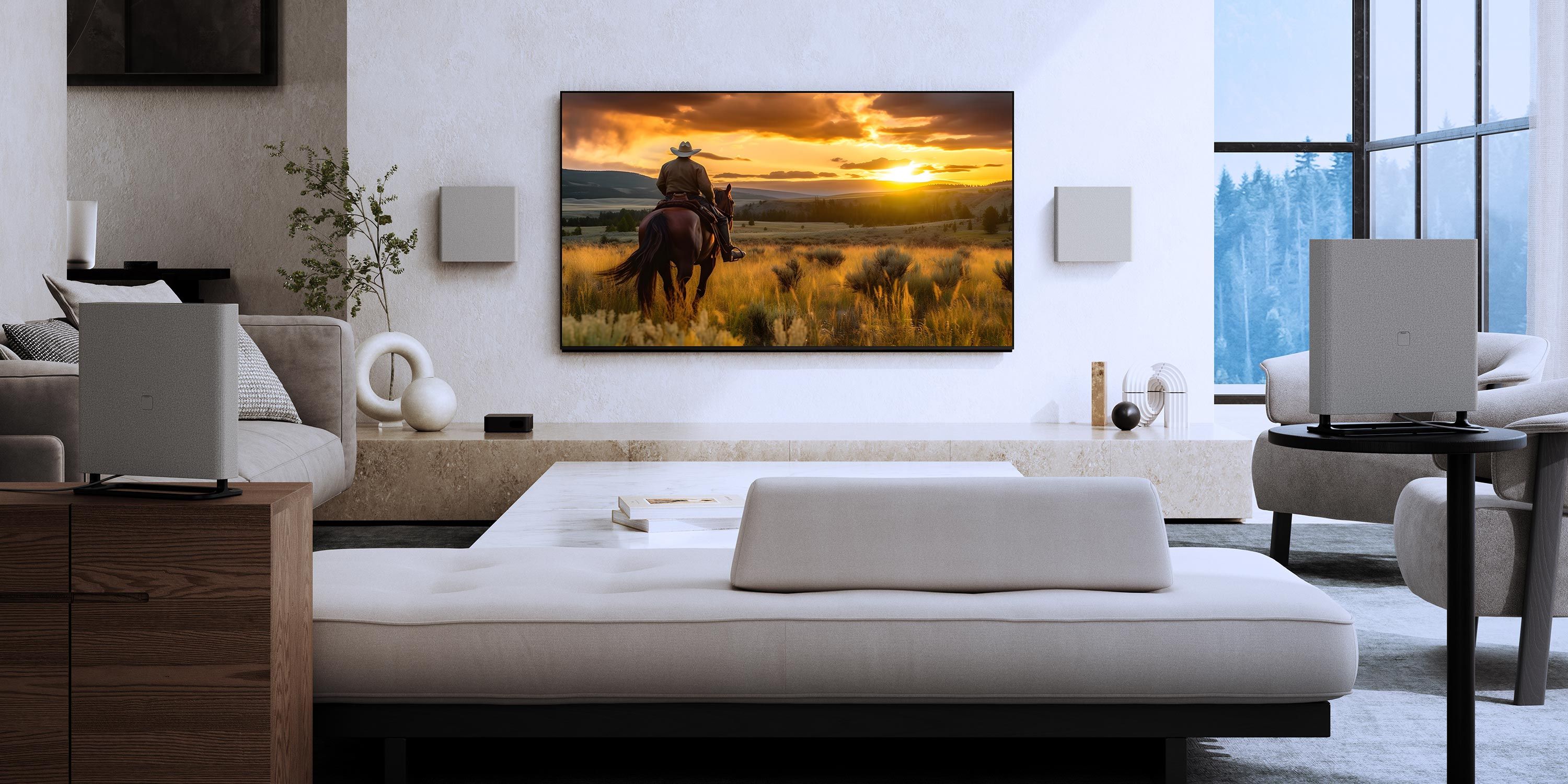 A modern media room with light furnishings, wall-mounted speakers, and a large TV showing a sunset scene over an open field.