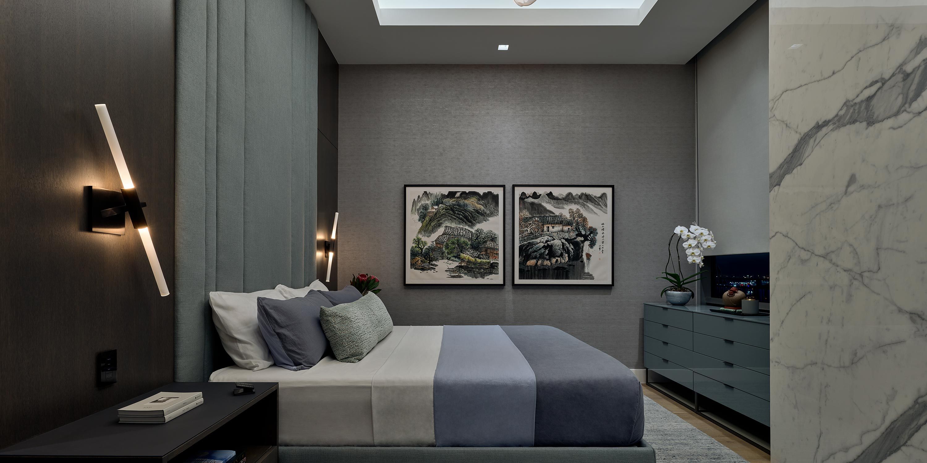 A cozy modern bedroom with a sleek wall sconce, a bed dressed in muted tones, and two framed artworks above the bed.