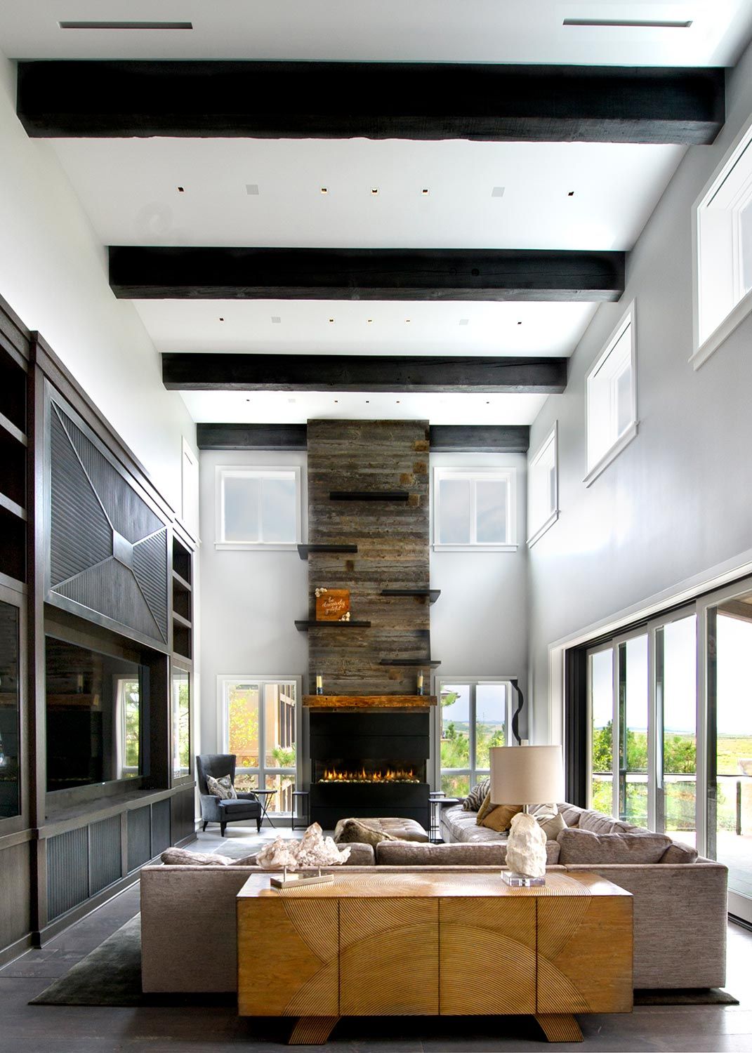 A spacious living room with high ceilings, exposed beams, a large stone fireplace, and comfortable seating, filled with natural light from the expansive windows.