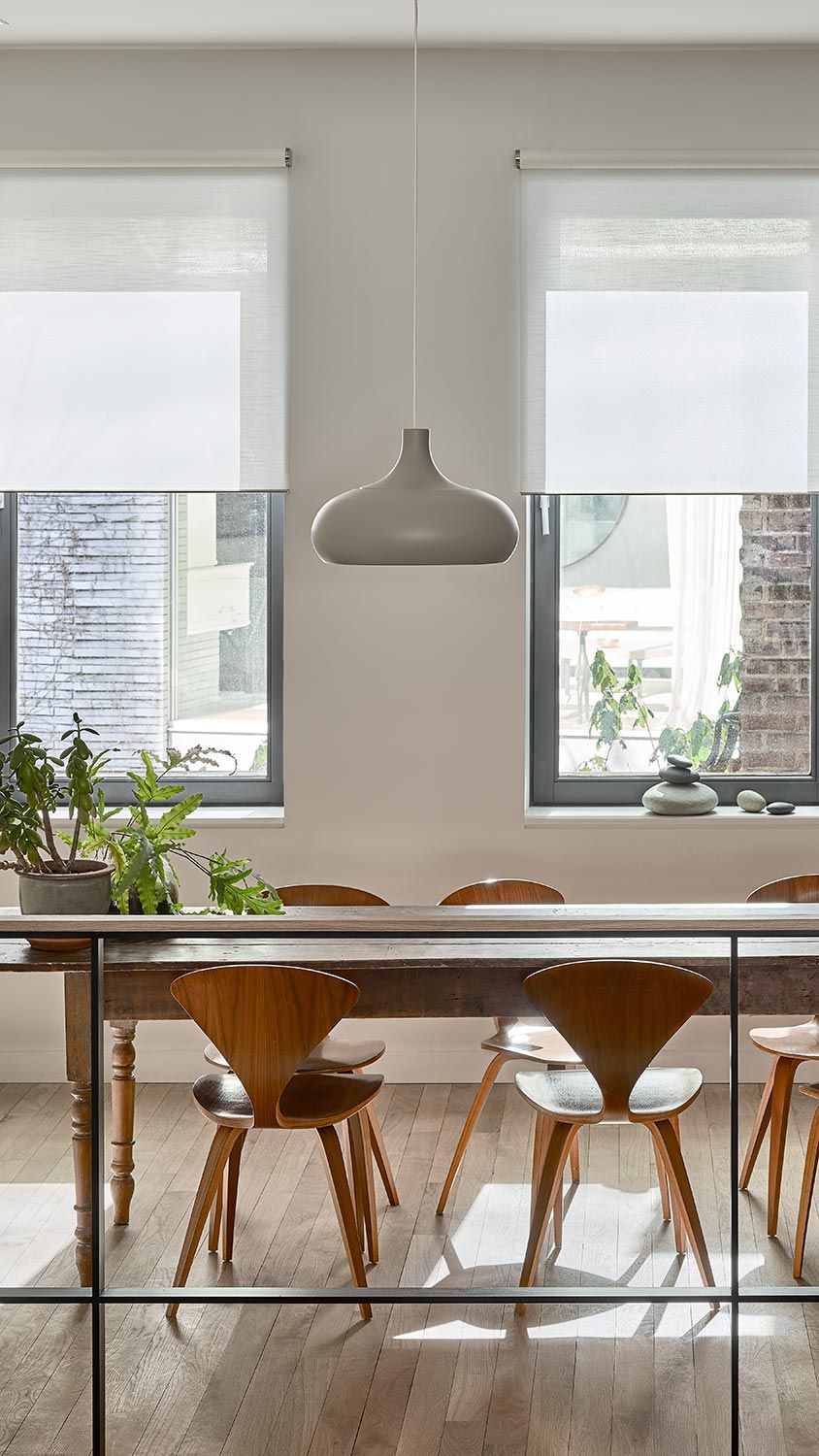 A modern dining area with large windows, stylish roller shades, and wooden dining chairs, illuminated by natural light.