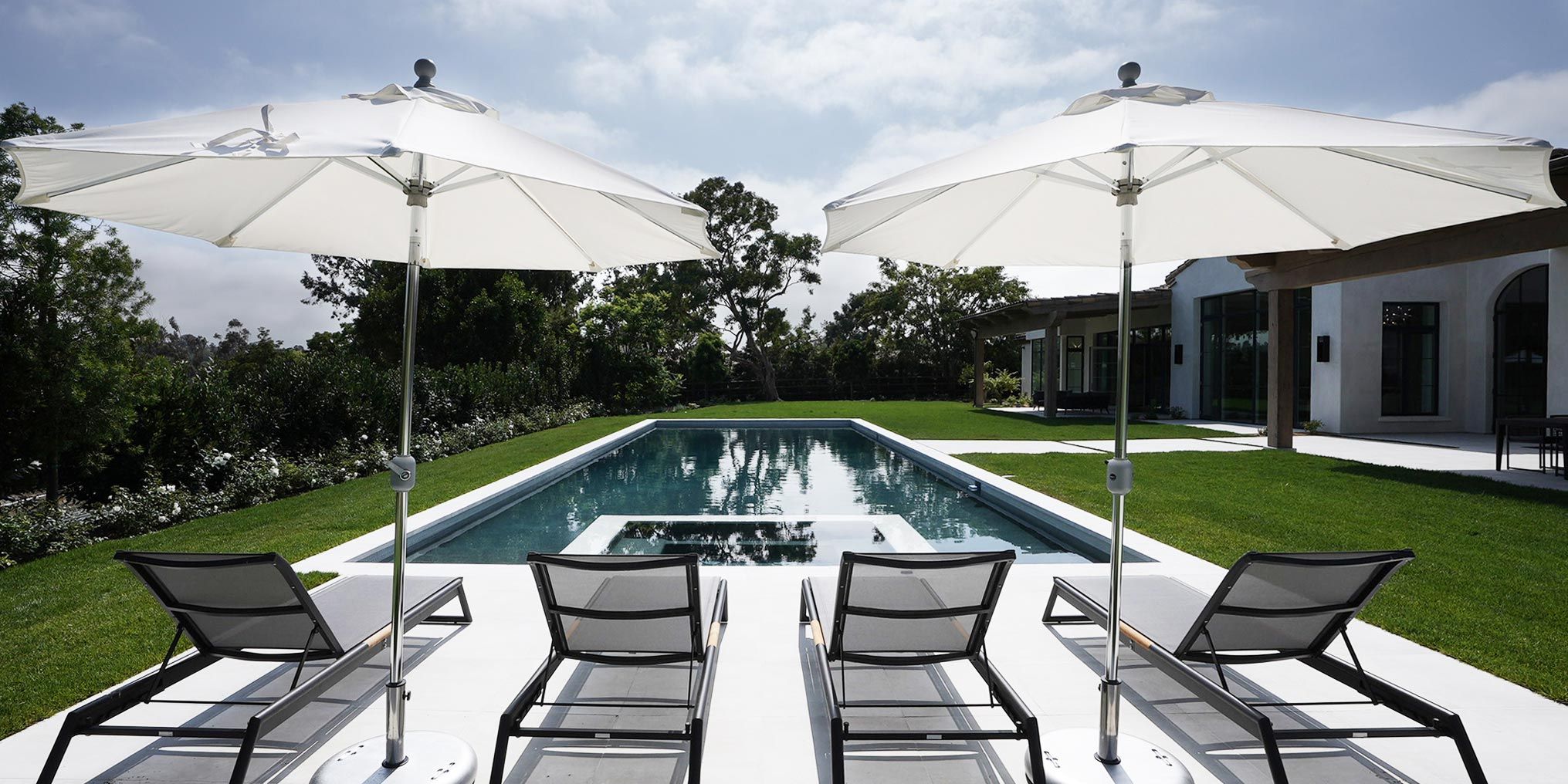 A serene pool area with two white umbrellas and sun loungers overlooking a sleek, modern pool surrounded by greenery.