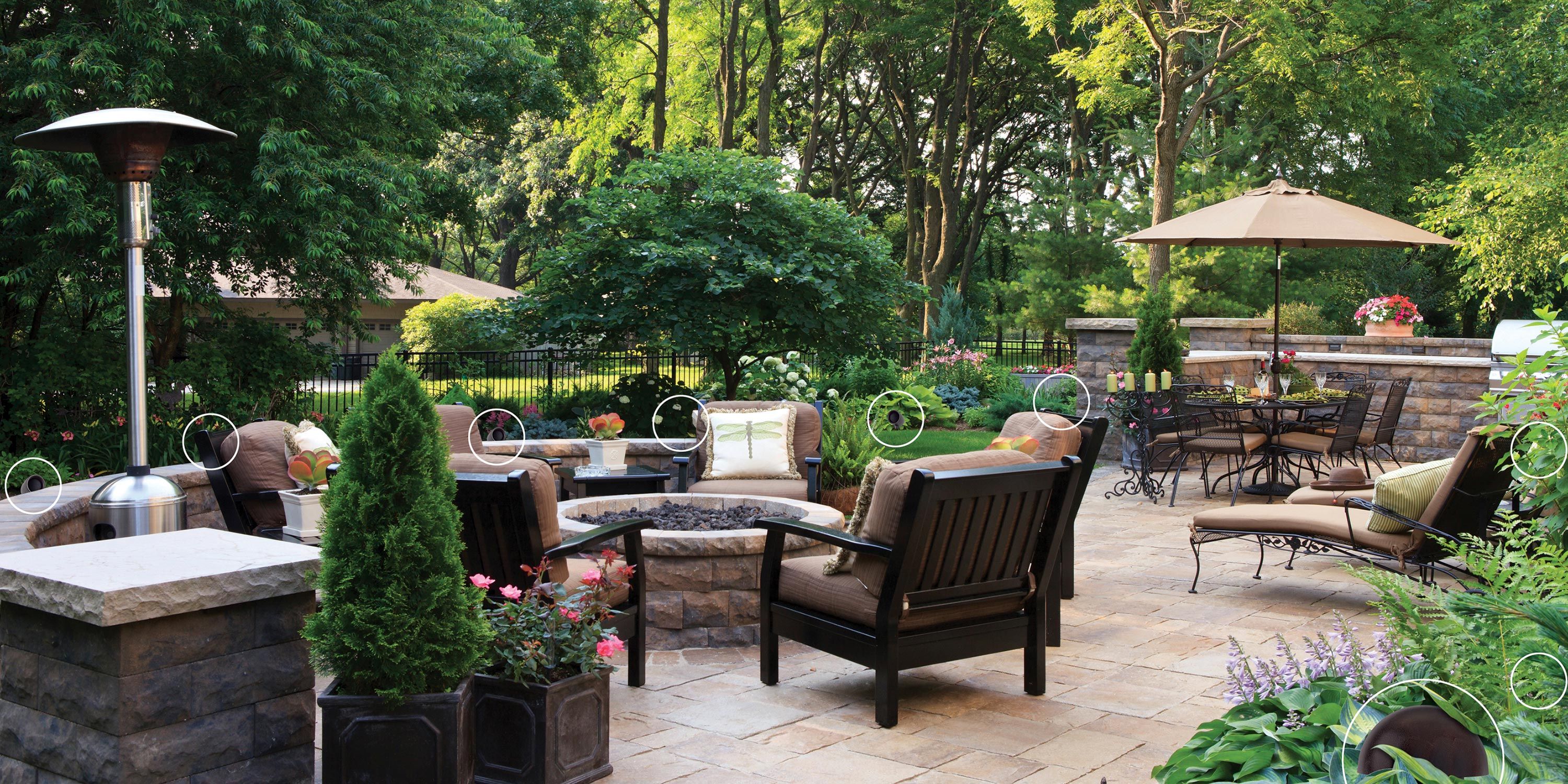 A beautifully landscaped backyard patio featuring comfortable seating around a fire pit, surrounded by lush greenery and outdoor dining space under an umbrella.