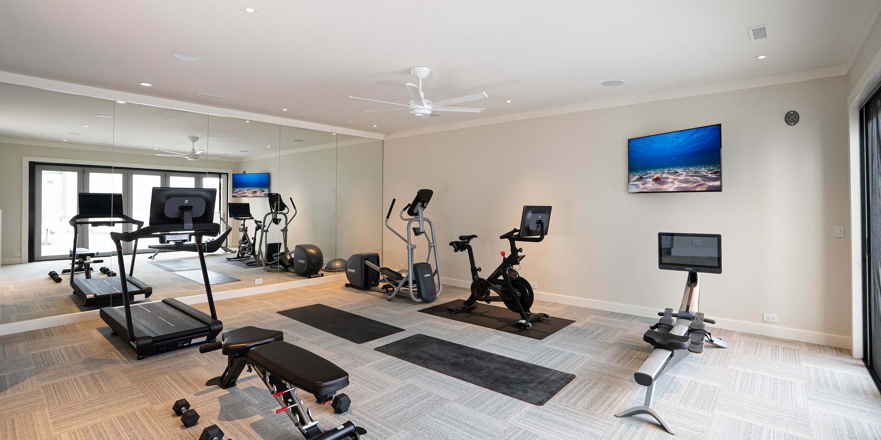 A well-lit home gym featuring cardio equipment, a mirrored wall, and a mounted TV displaying an underwater scene.