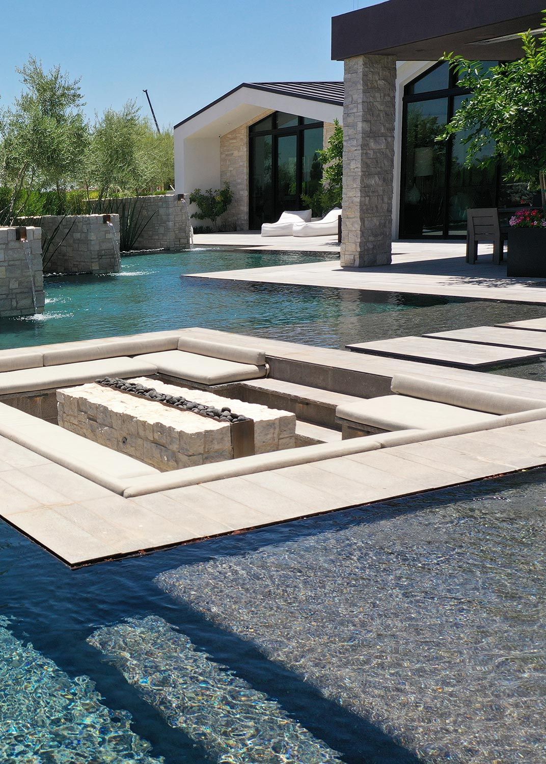 A modern outdoor living space featuring a sunken seating area with a firepit, surrounded by a reflective pool and stone accents, leading to a stylish house with large windows.