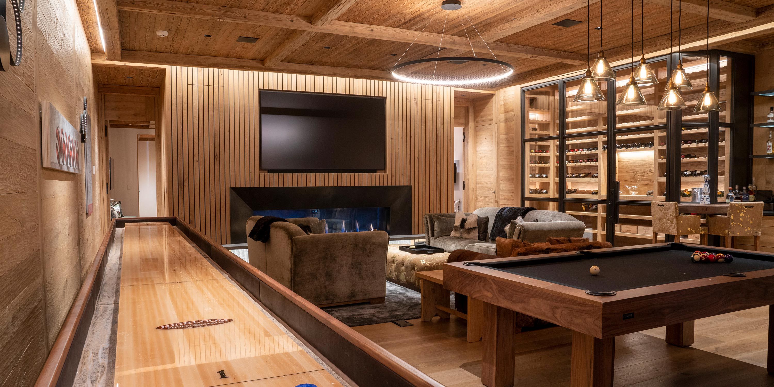 A luxurious game room with wooden walls, a shuffleboard table, pool table, and a seating area near a fireplace, alongside a wine storage display.