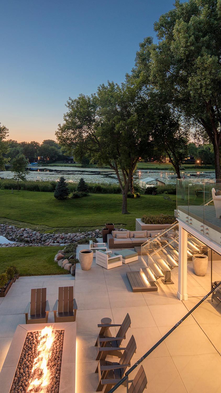 A spacious outdoor patio with a firepit, comfortable seating, and scenic views of a lake and greenery at sunset.