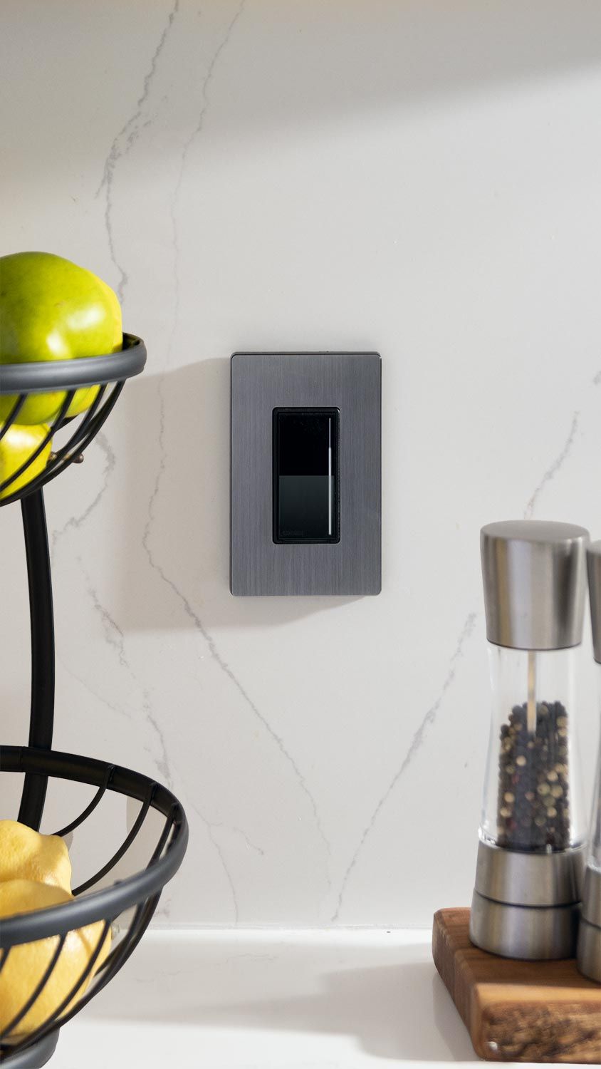 A sleek wall control switch is mounted on a kitchen backsplash next to a fruit basket and pepper grinder.